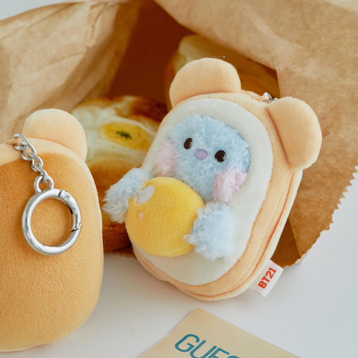 Two BT21 plush keychains in a paper bag. One keychain features a light blue plush character holding a yellow sphere in a toast-shaped frame. The other keychain is a tan, rounded rectangle.