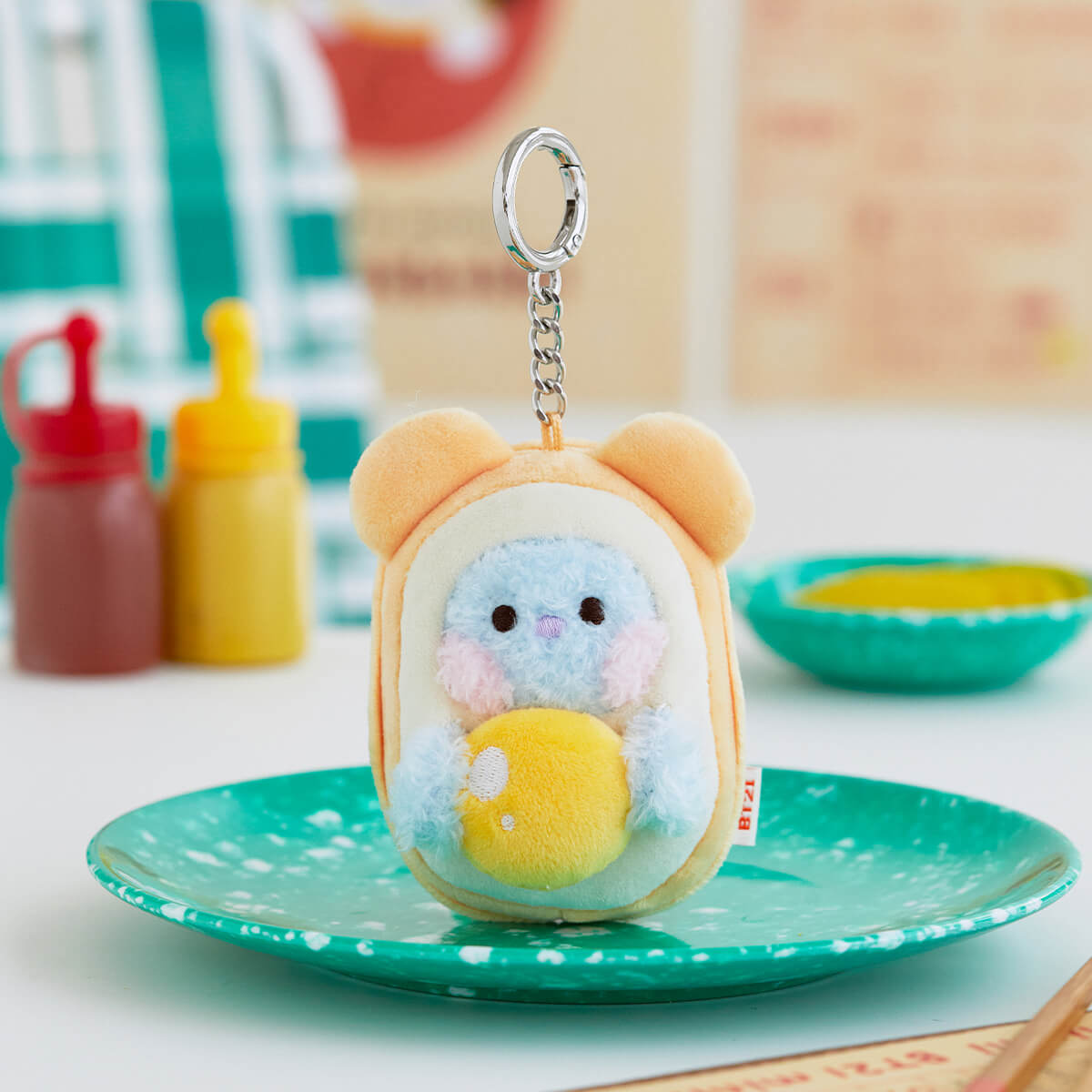 A blue and yellow BT21 character plush keychain with a silver clip, resting on a green speckled plate.