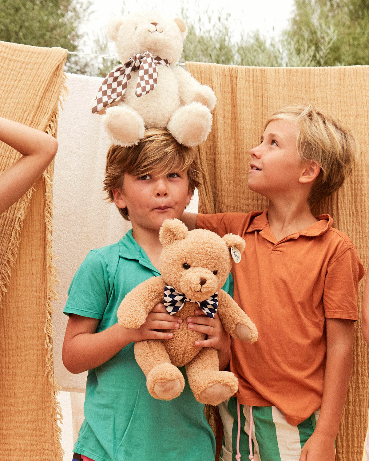 Two boys holding teddy bears in front of hanging blankets.