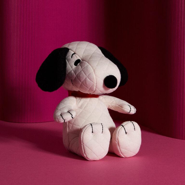 White quilted Snoopy plushie with black ears and nose, sitting against a pink background.