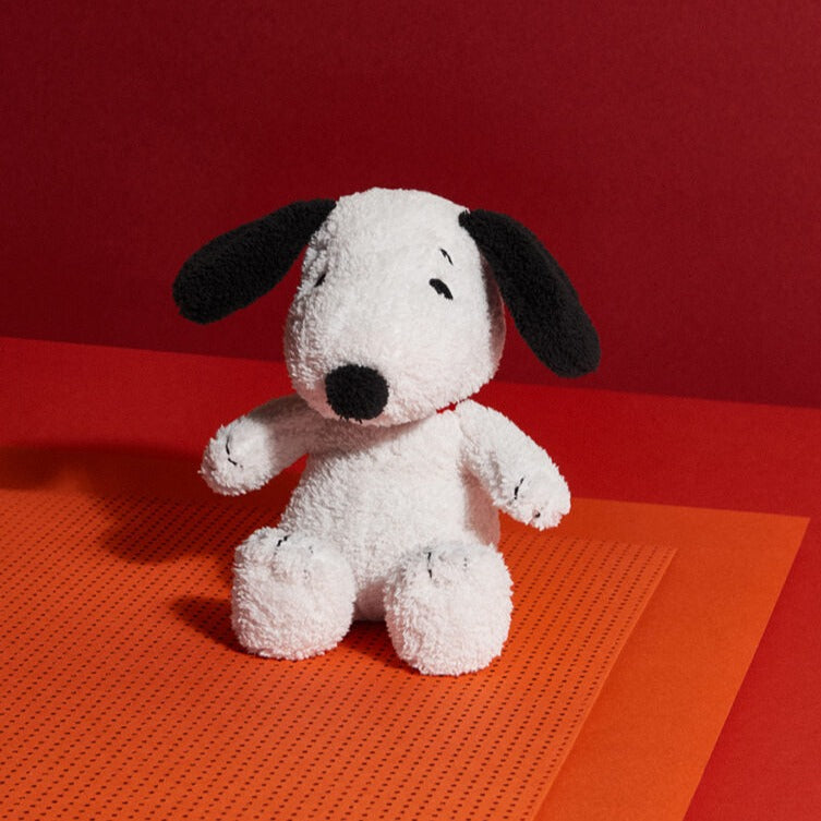 A white Snoopy plush doll with black ears and nose sits on orange papers against a red backdrop.