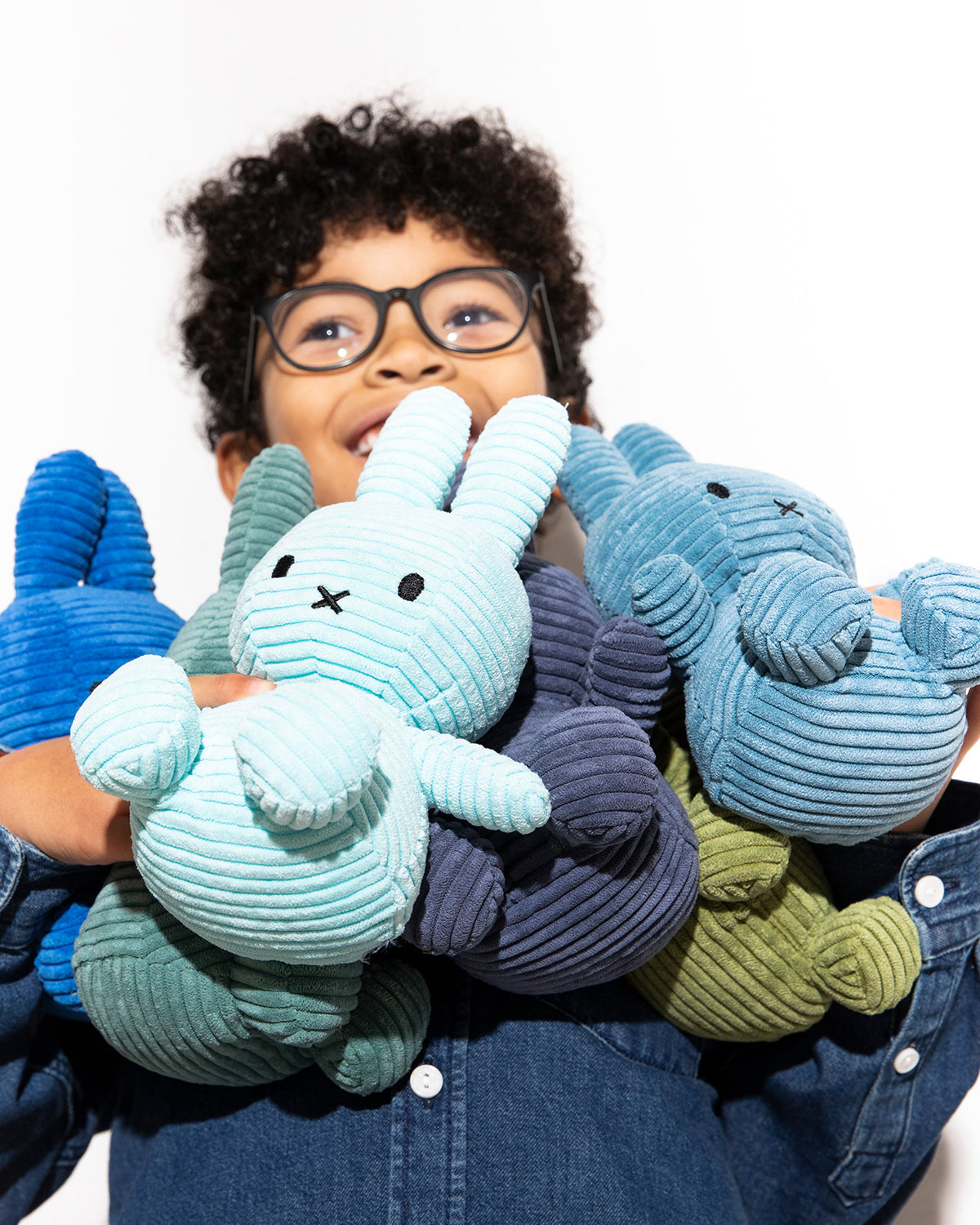 Young boy with curly hair and glasses holding several corduroy bunny stuffed animals.