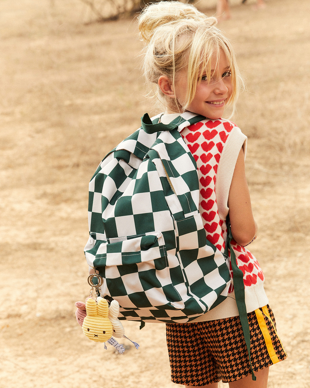 Child wearing a green and white checkered backpack with a heart-patterned shirt and plaid shorts.