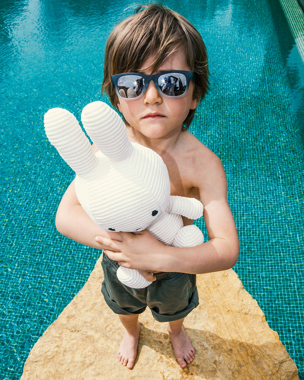 A young boy in sunglasses holding a stuffed rabbit by a pool.