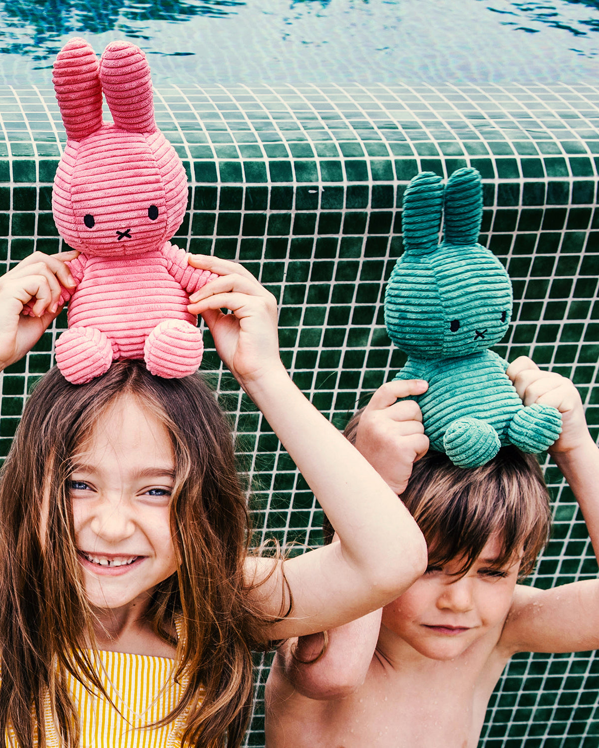 Two children holding pink and green plush toys by a pool.