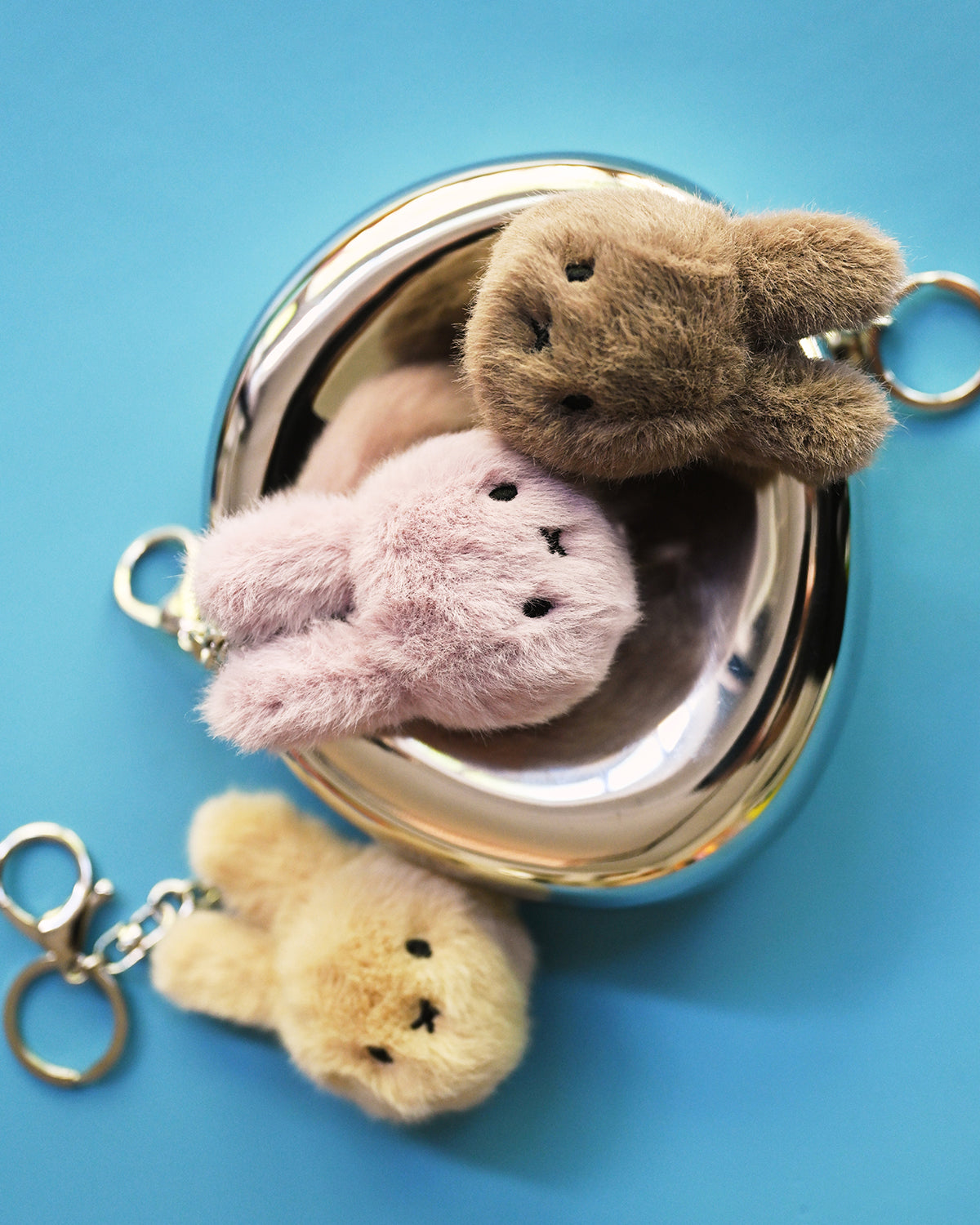 Three fluffy bunny keychains in a shiny, round bowl on a blue background.