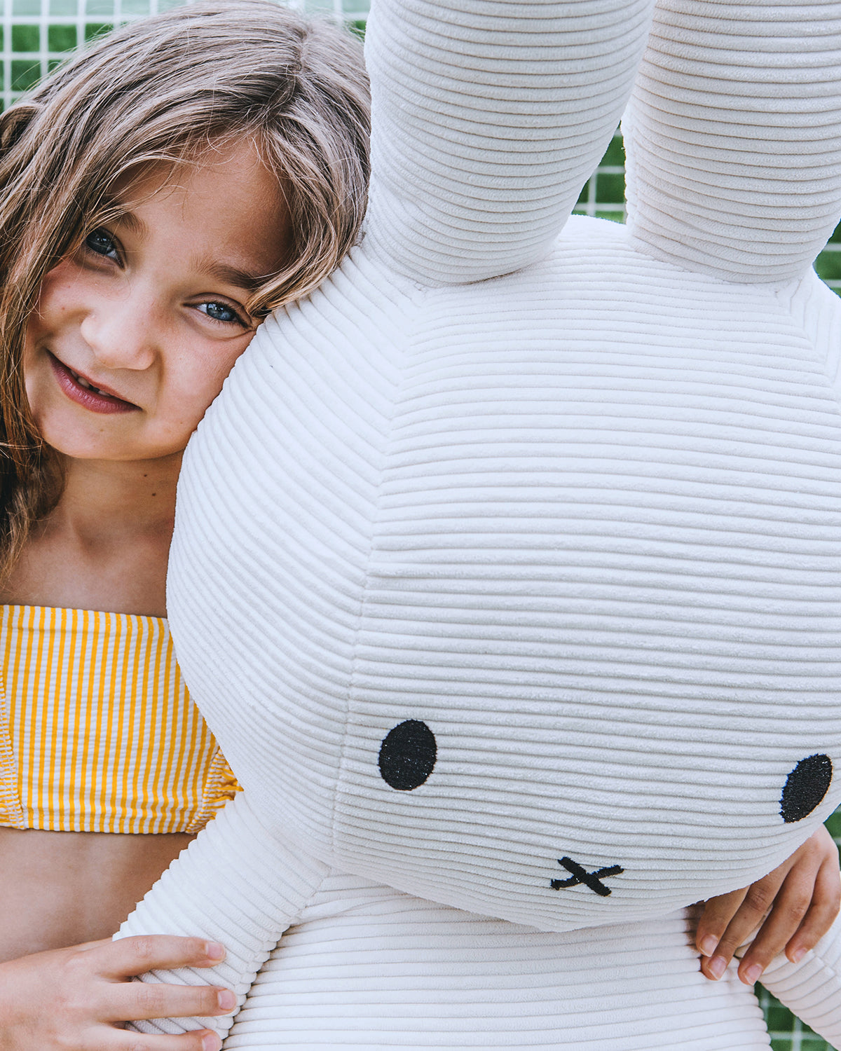 Young girl hugging a large Miffy plush doll. The girl wears a yellow and white striped top.