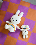 Two plush toy rabbits, one larger than the other, lie on a checkered purple and orange surface. Both are white with stitched facial features and rainbow detailing.