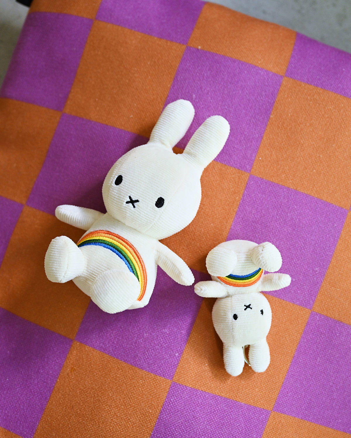 Two white stuffed rabbits with rainbow accents on a purple and orange checkered background.