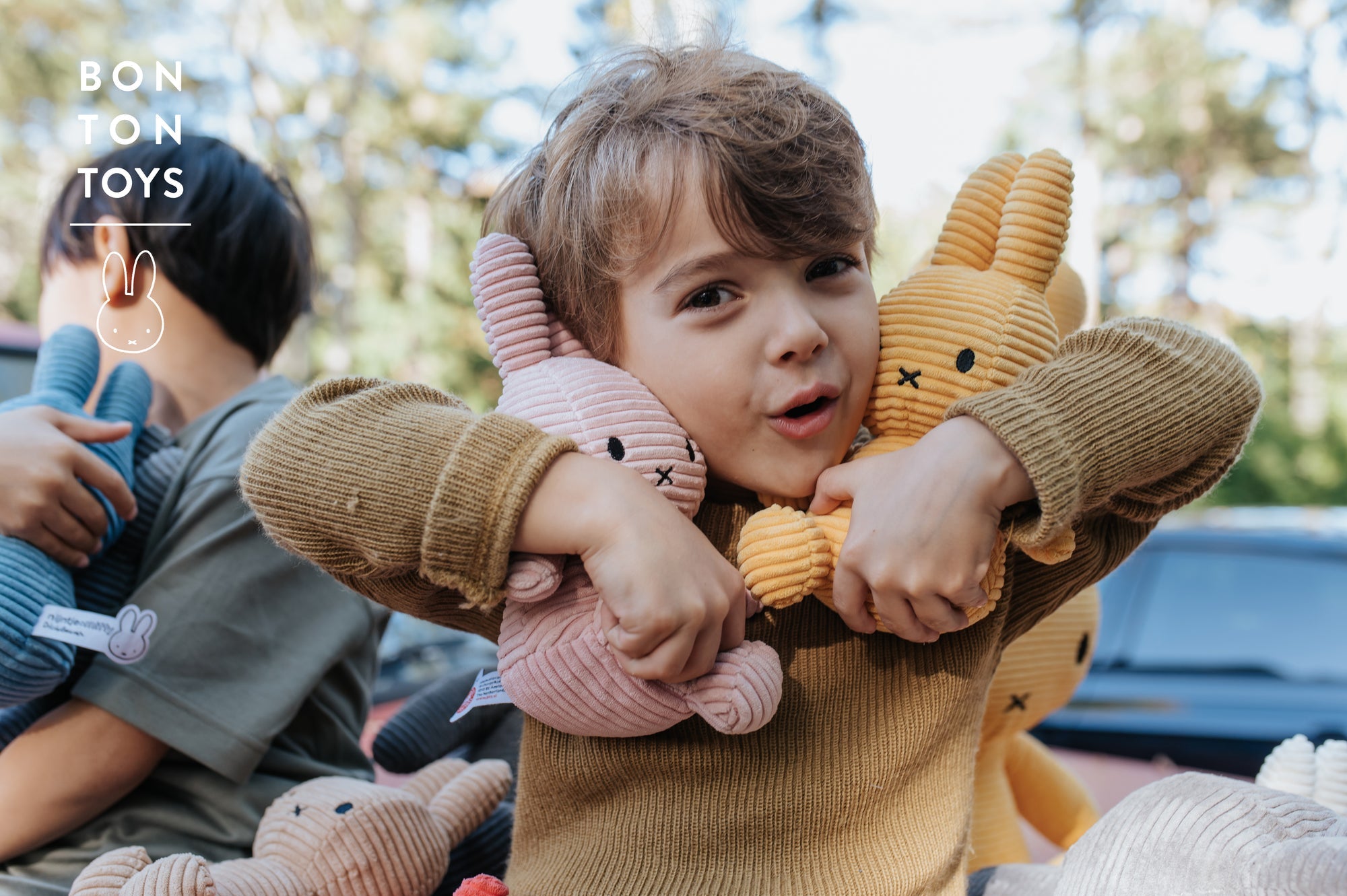 Child hugging pink and yellow corduroy bunny plushes, tan sweater, outdoor setting.