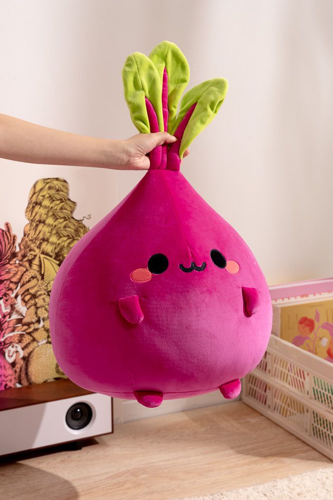 A bright pink plush beet toy with green leaves is held up by a person in front of a beige wall.