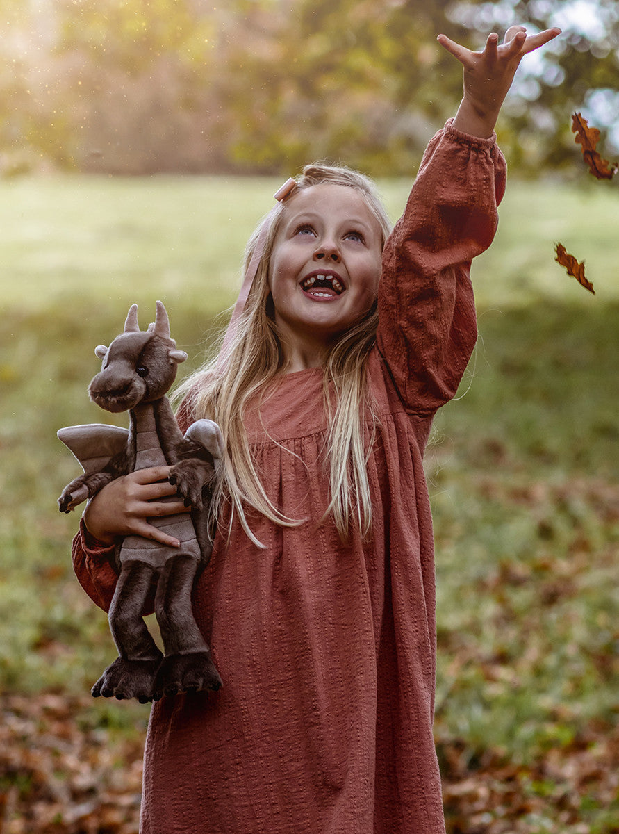 Young girl in a pink dress holding a dragon toy outdoors with leaves on the ground