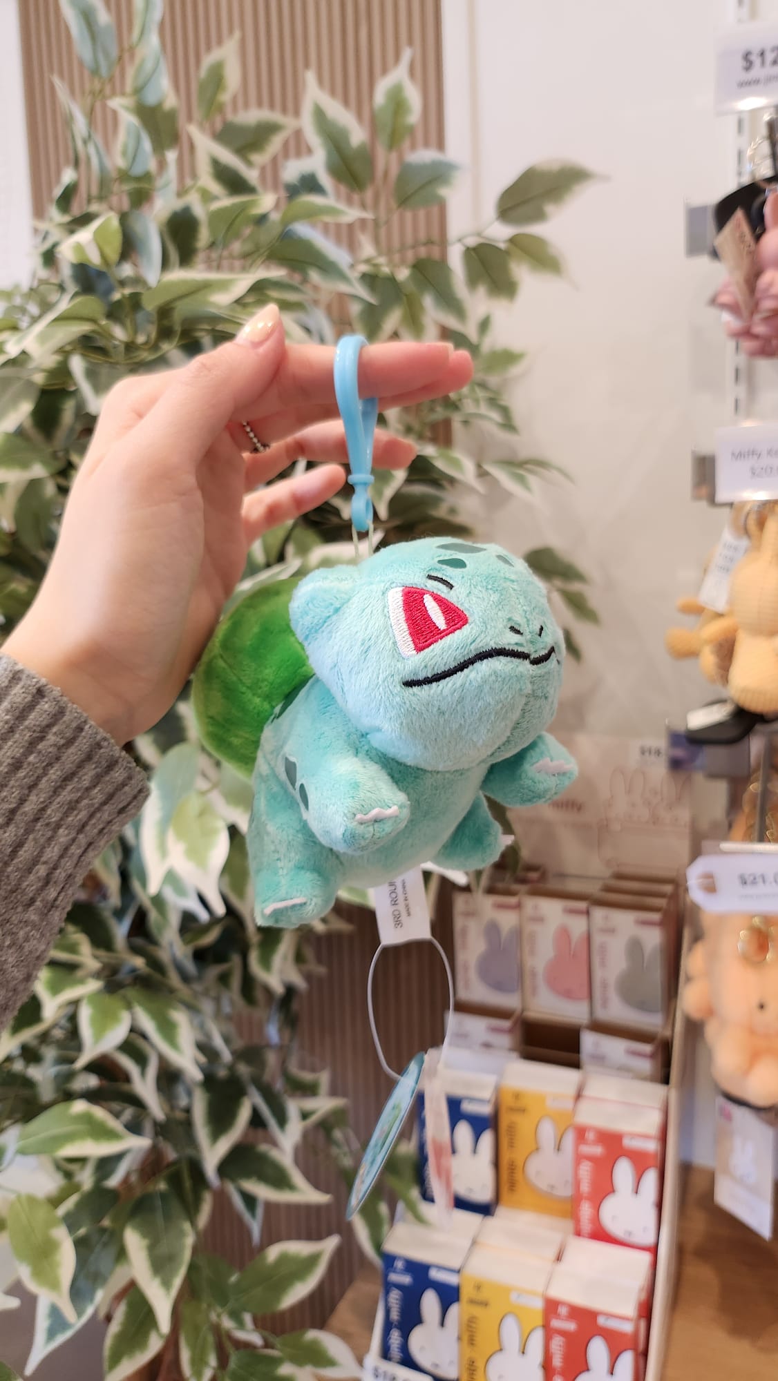 Hand holding a teal Bulbasaur plush with a blue clip, with merchandise in the background.