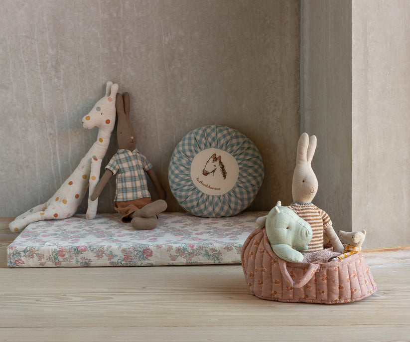 Stuffed animal toys: giraffe, bunny in plaid shirt, gingham pillow, bunny in striped shirt, hippo, and mouse, in a pink floral quilted basket on a wooden floor.
