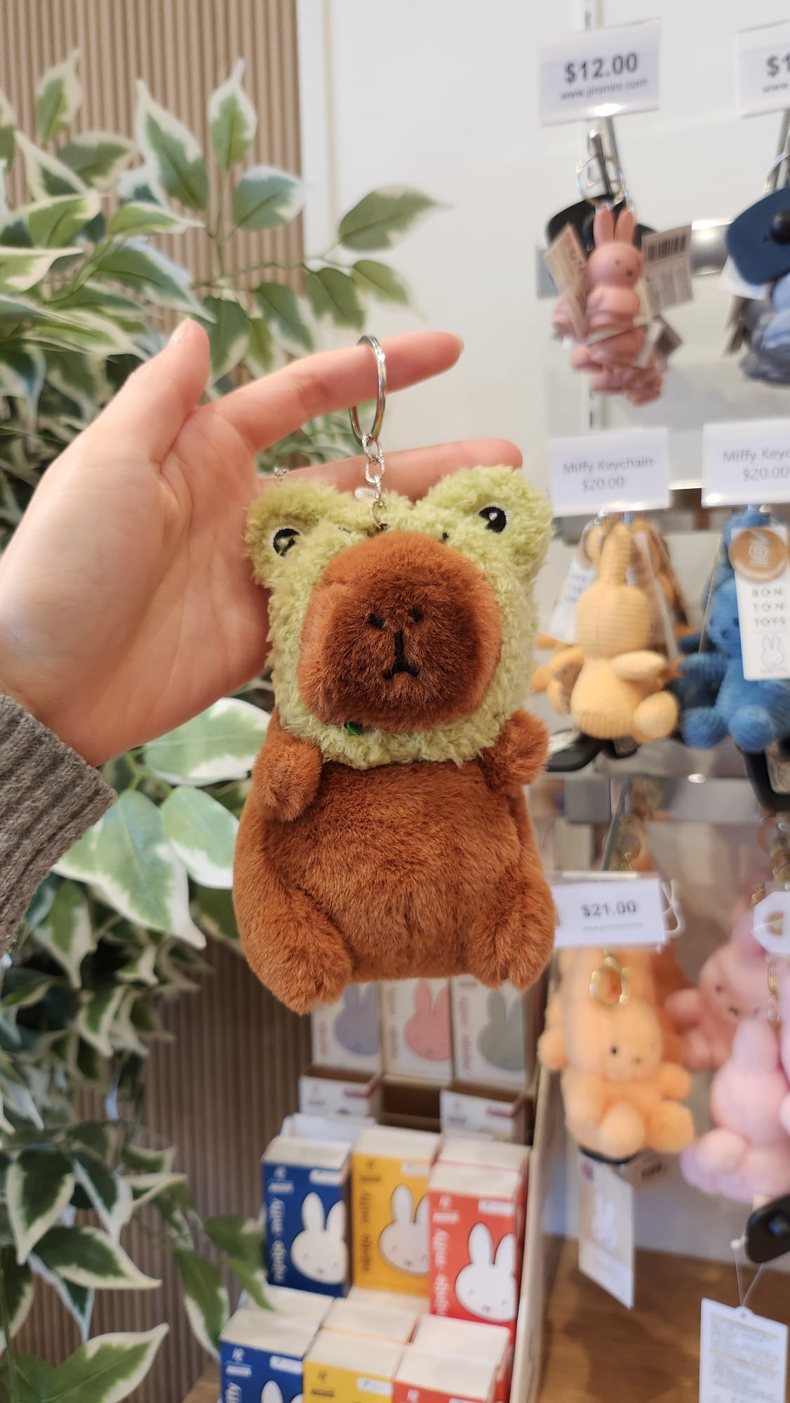 Close-up of a brown and green plush keychain of a bear-like creature being held up in front of a shelf of Miffy products.