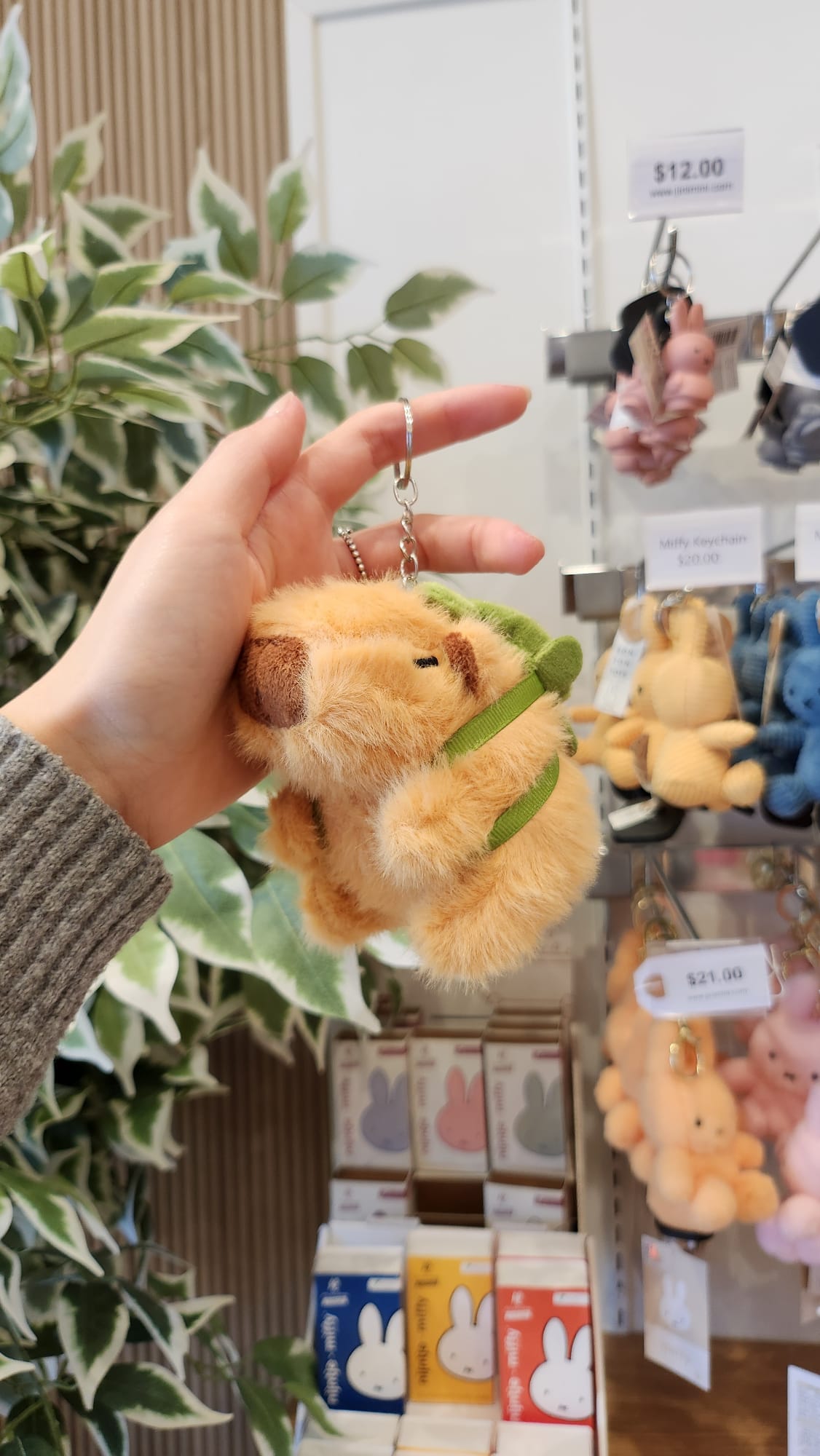Hand holding a tan, fuzzy bear keychain with a green strap, displayed in a store.