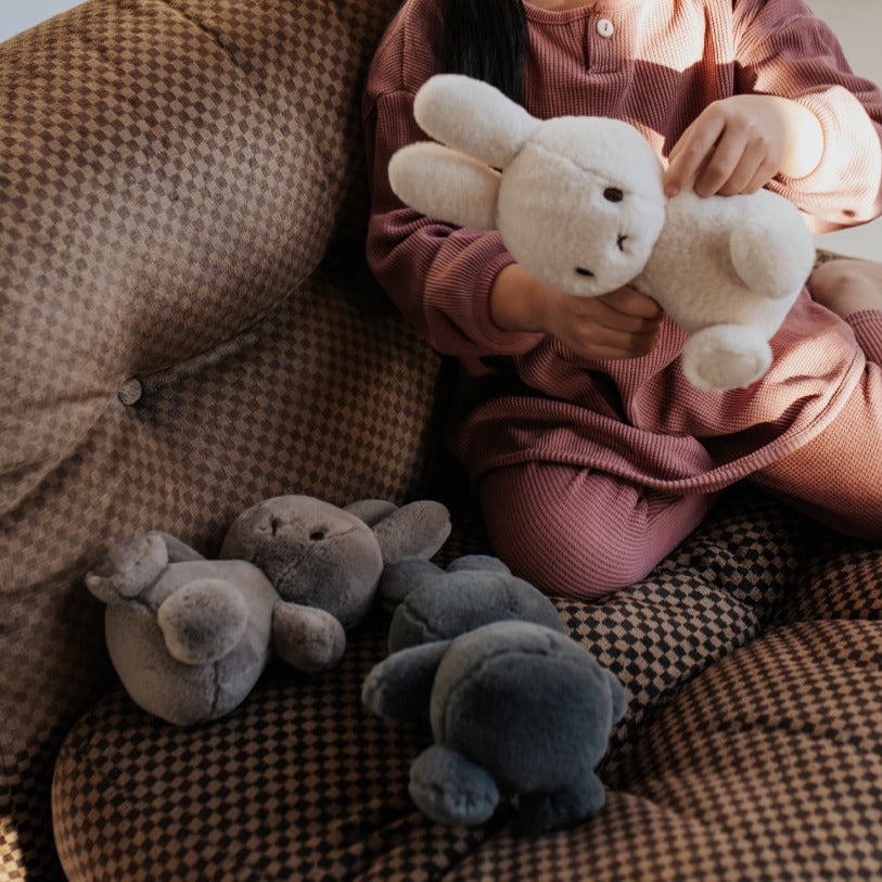 Child in rose-colored clothing sitting on a brown patterned couch while holding a white plush rabbit and surrounded by gray plush toys.