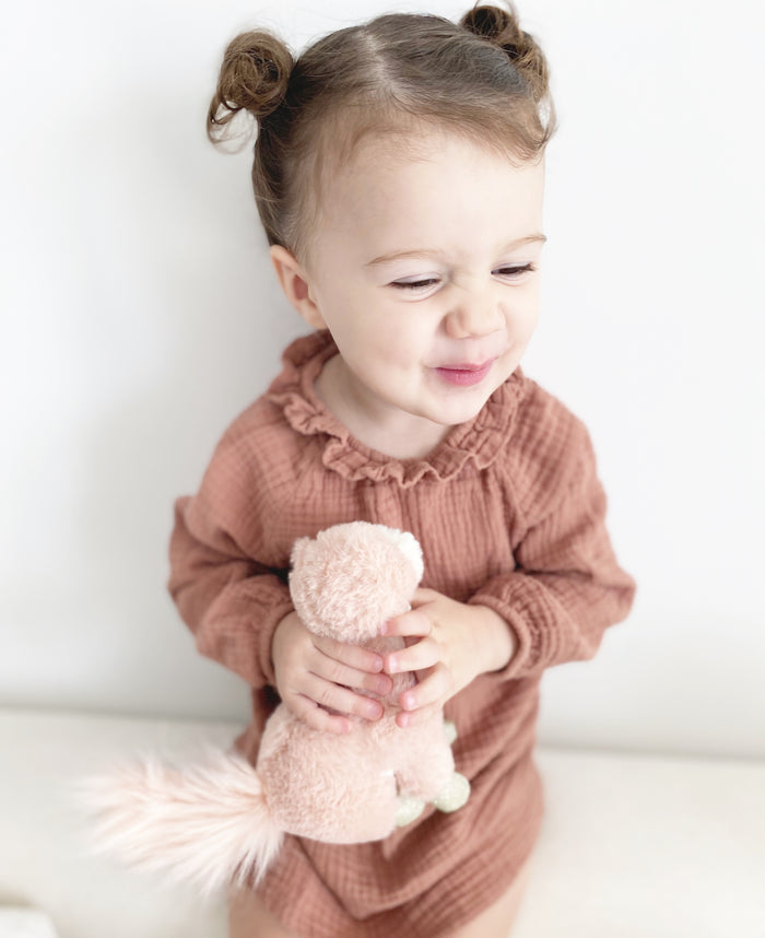 A young child with pigtails wears a pink ruffled romper and holds a stuffed squirrel toy.