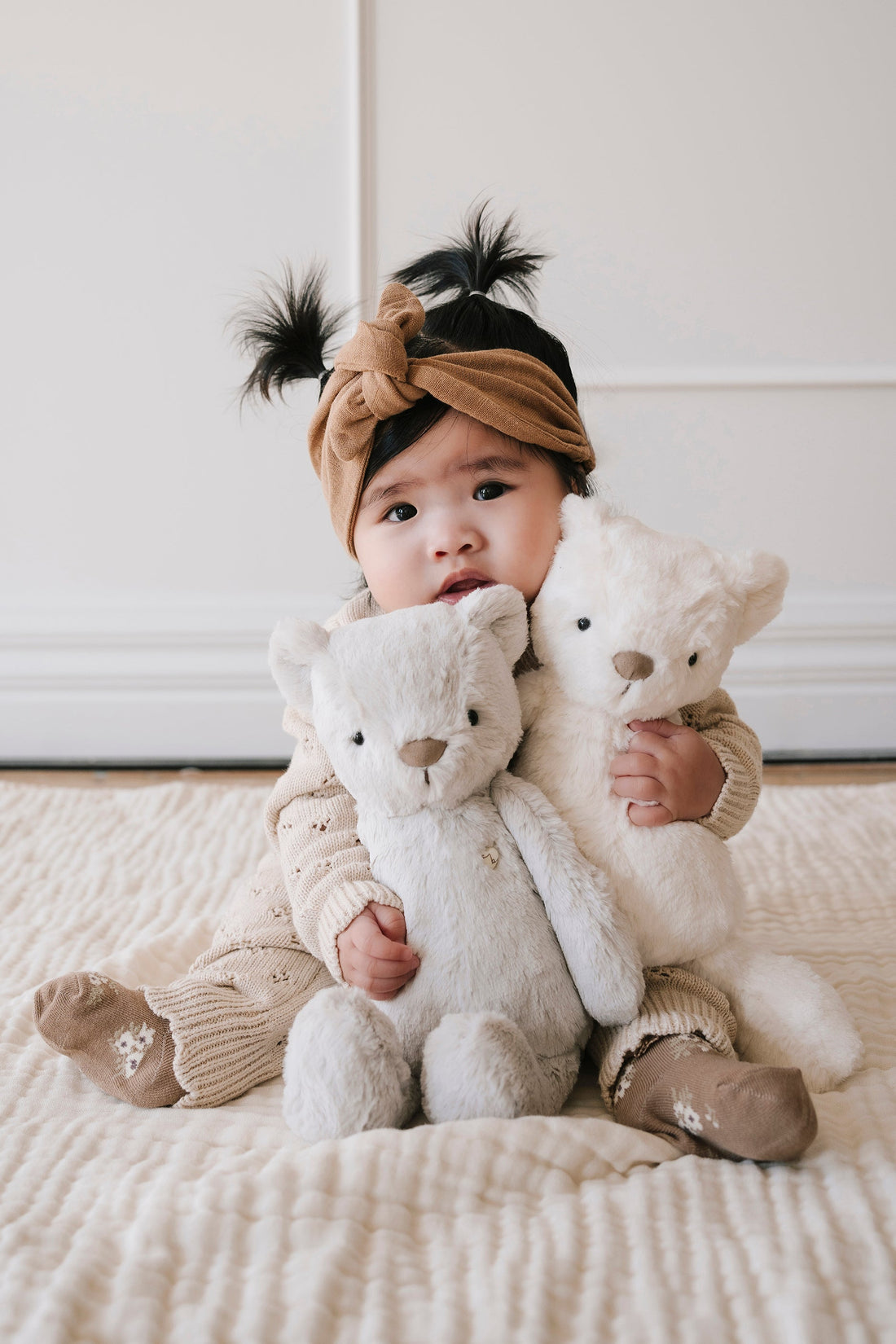 A baby wearing a brown headband and holding two teddy bears.