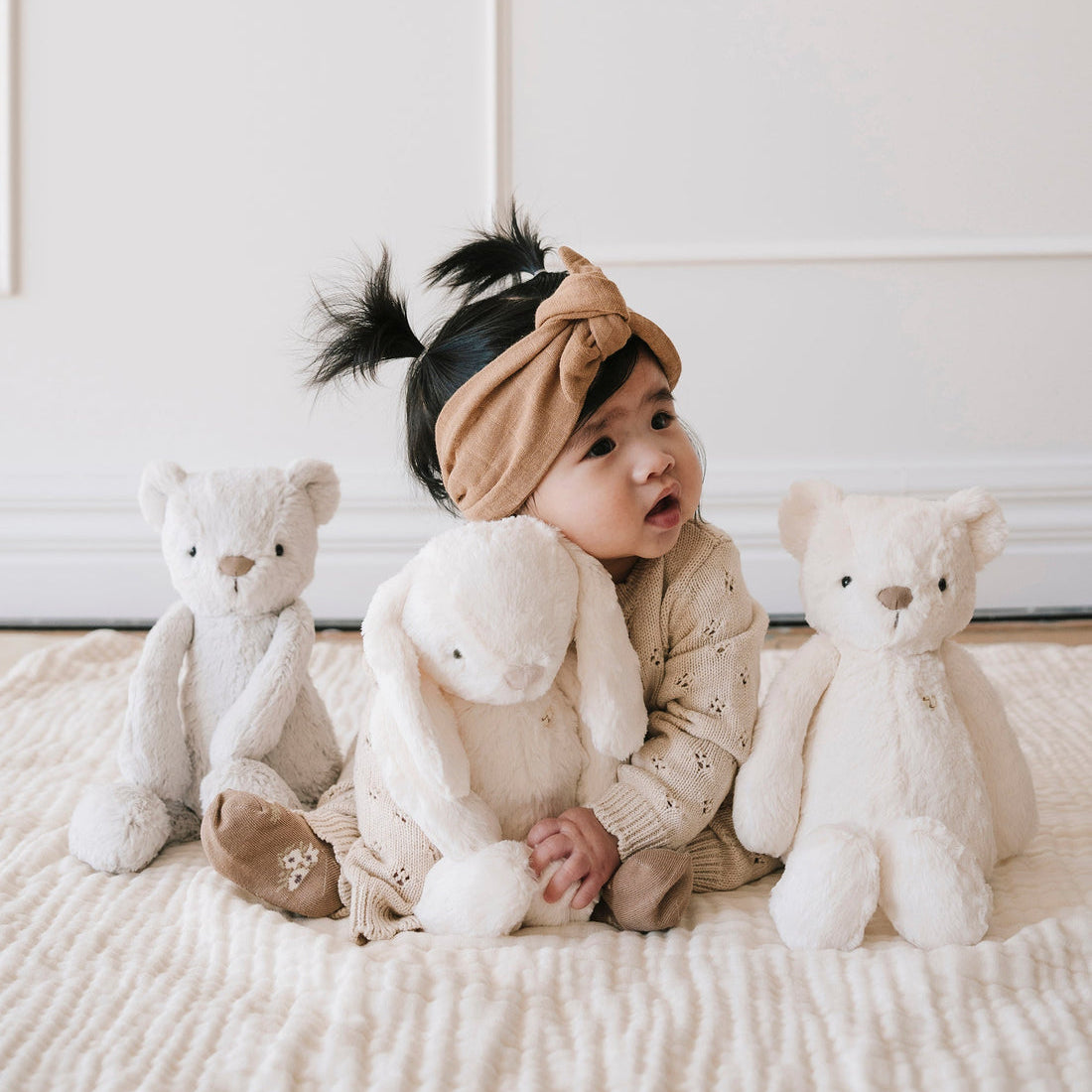 Baby girl in beige outfit with stuffed animals.