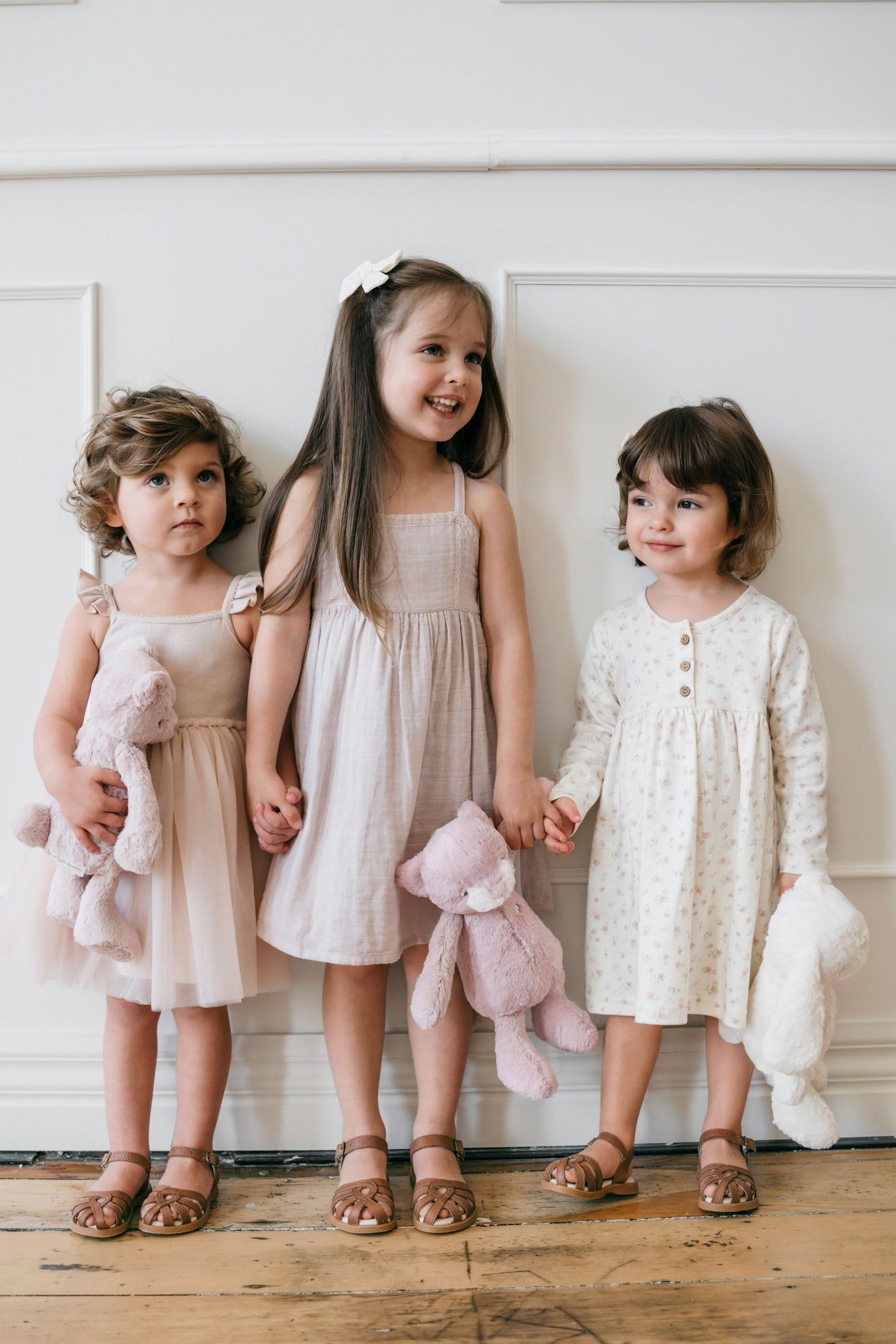 Three young girls in dresses and sandals, holding stuffed animals.
