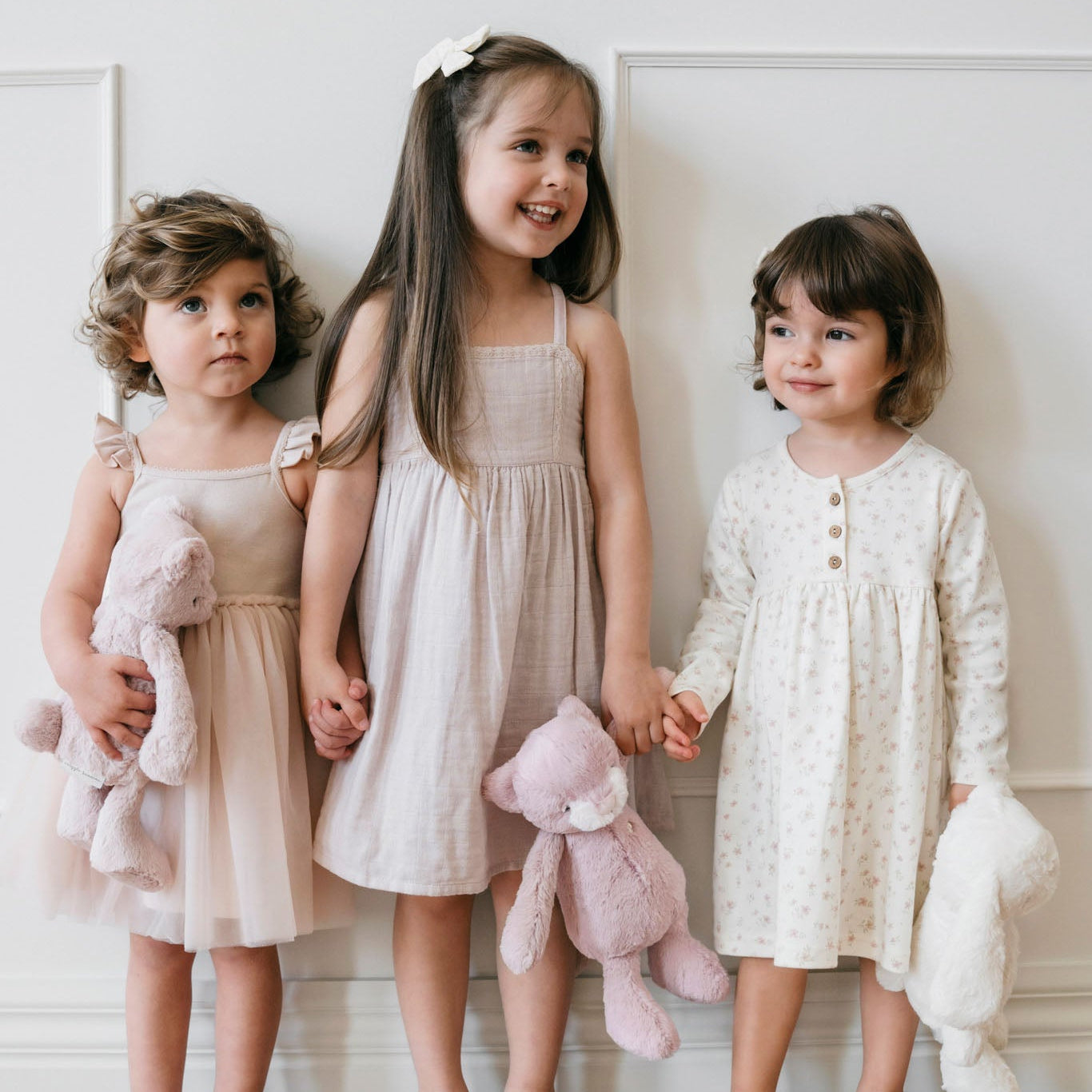 Three young girls in dresses and sandals, holding stuffed animals.