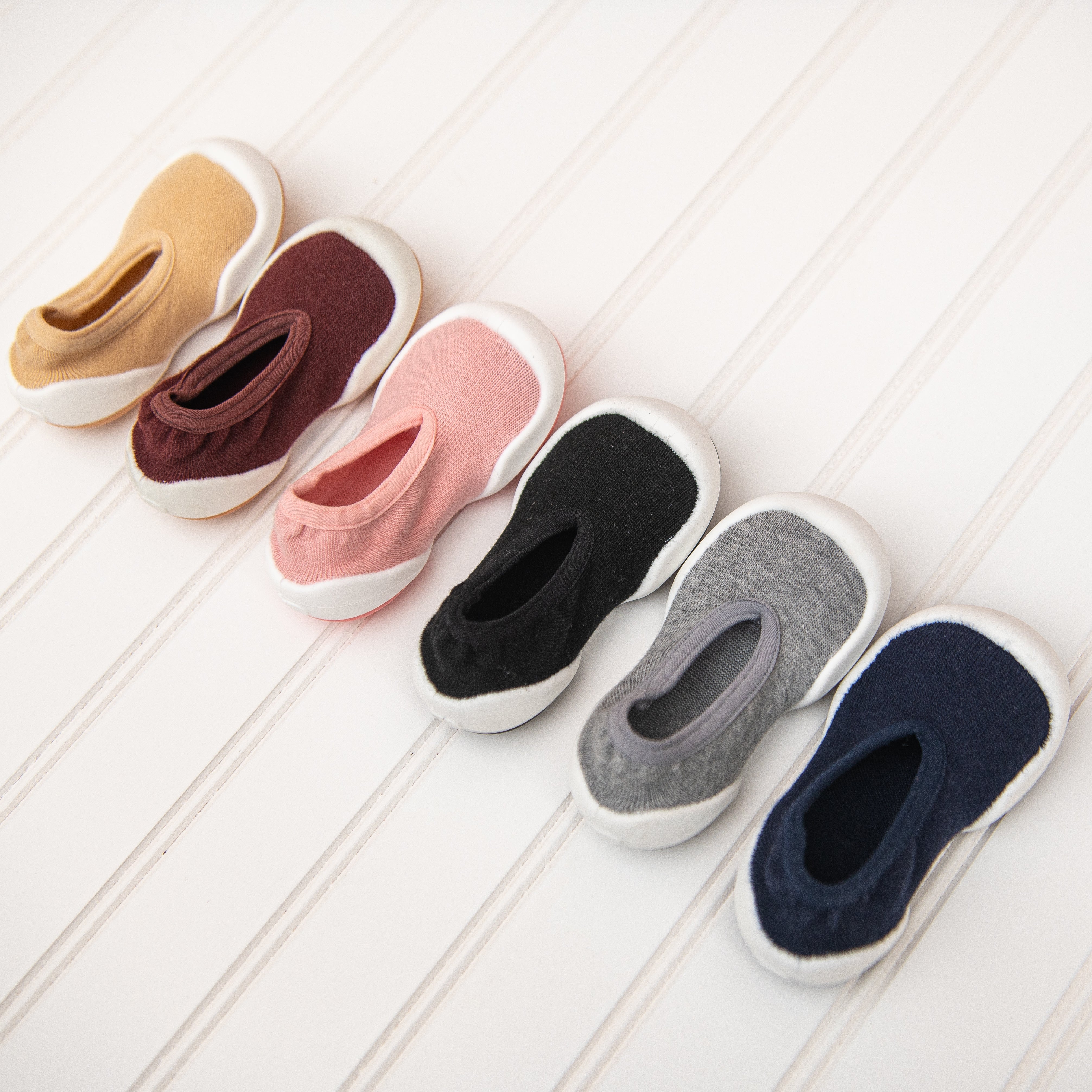Six baby sock-shoes in a row on white wood. Colors include tan, maroon, pink, black, gray, and navy.