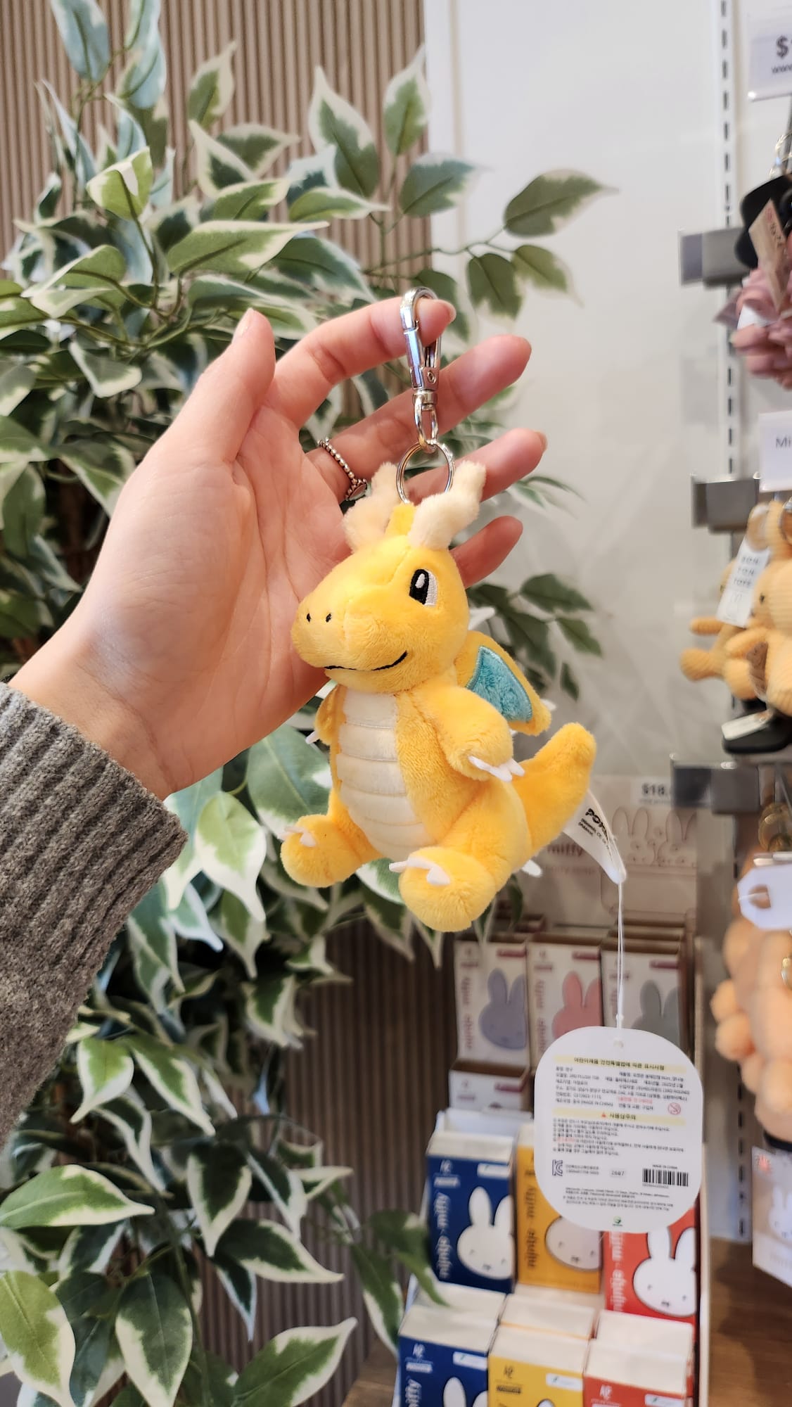 A plush yellow Dragonite keychain held in someone's hand with Miffy-themed products and an artificial green plant in the background.