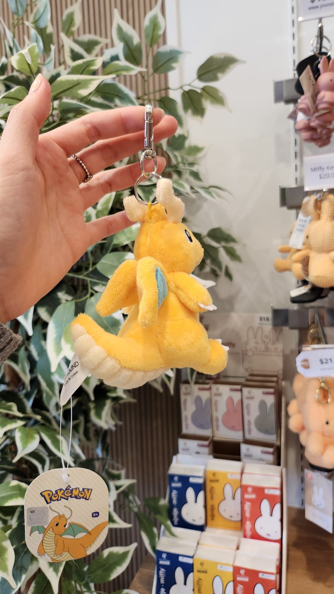 Close-up of a hand holding a yellow Dragonite Pokemon plush keychain in a store with Miffy merchandise in the background.