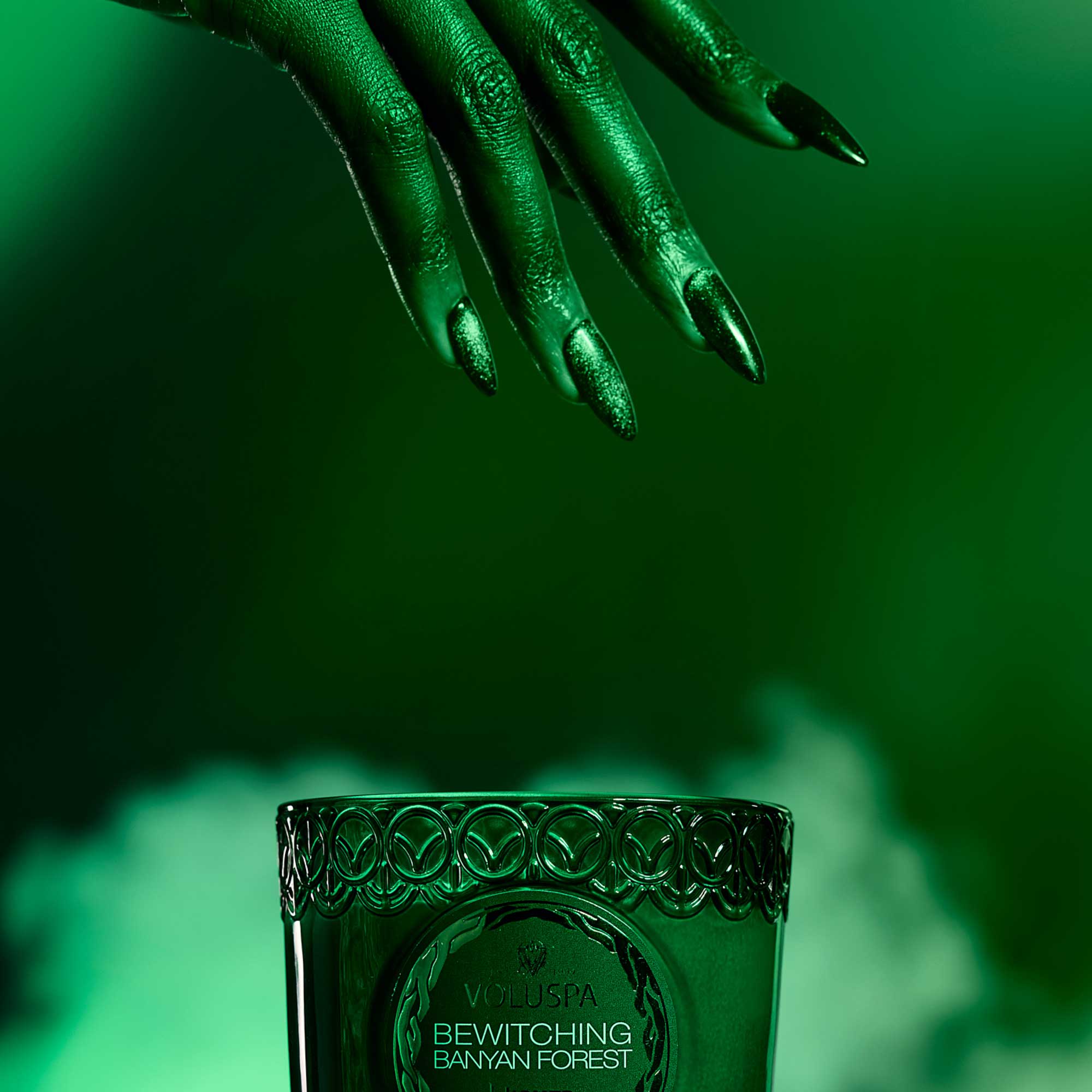 Green candle with intricate design and 'Bewitching Banyan Forest' text, hand with green nail polish above on a green background.