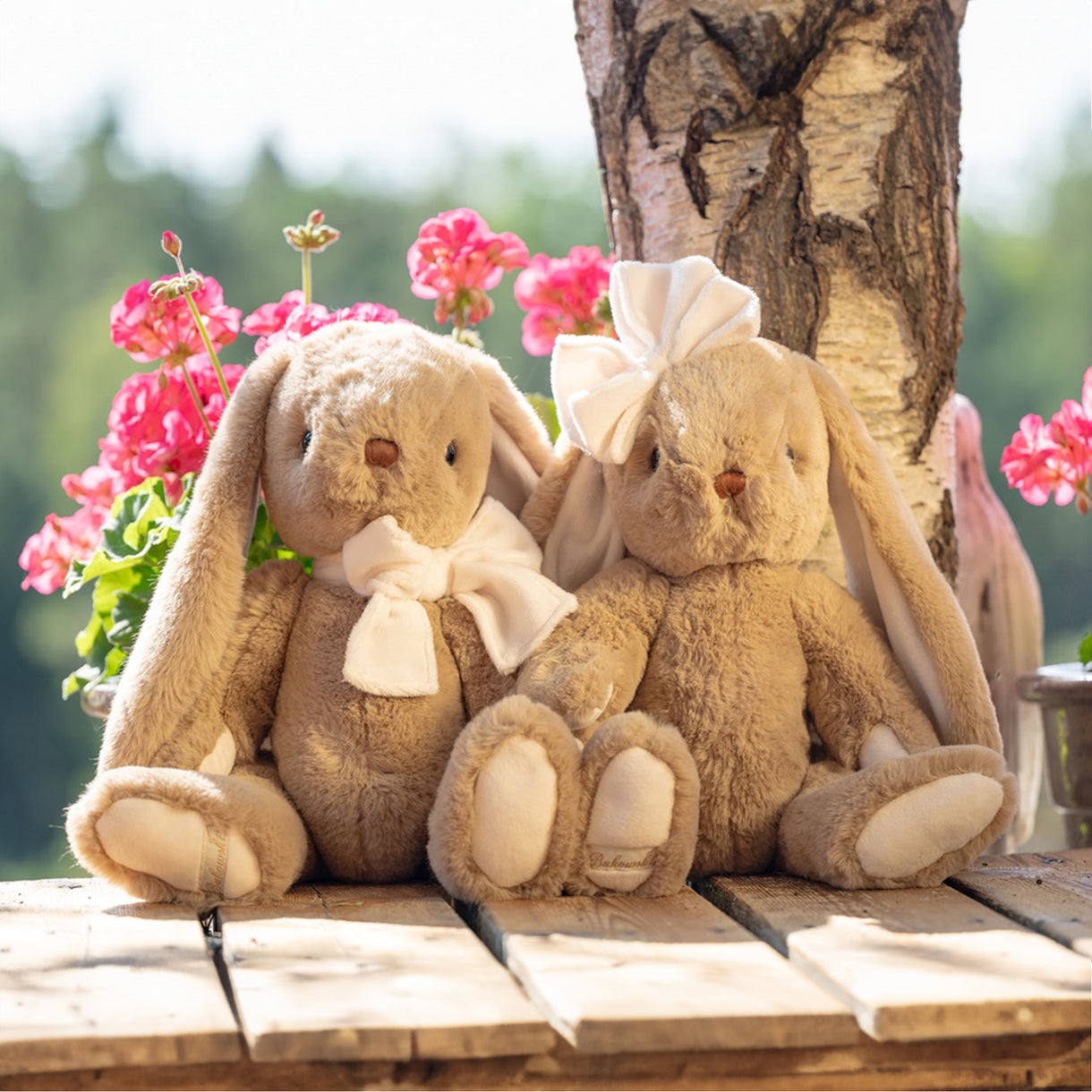Two plush bunnies with bows sitting on a wooden surface with flowers and a tree in the background.