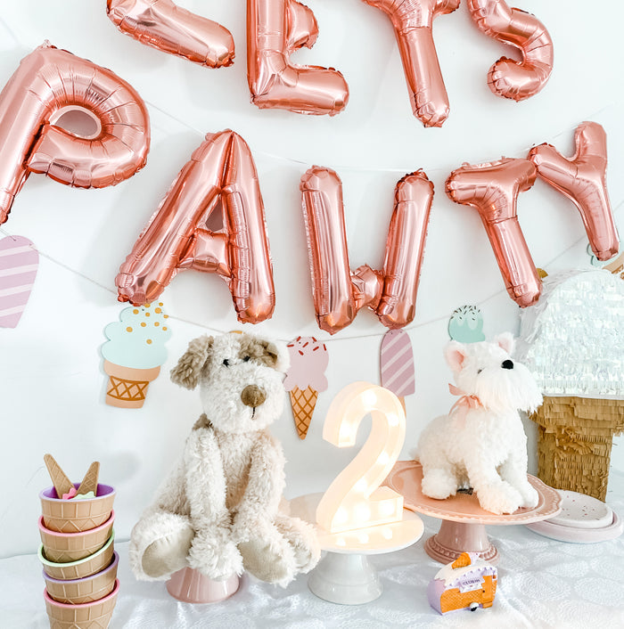 Festive birthday party setup with rose gold "Paws Party" balloons, a plush brown and white dog on a pink pedestal, a plush white dog with a pink bow on a pink plate, a light-up number "2" on a white pedestal, a stack of ice cream bowls, ice cream cone decorations, and a large ice cream cone pinata.
