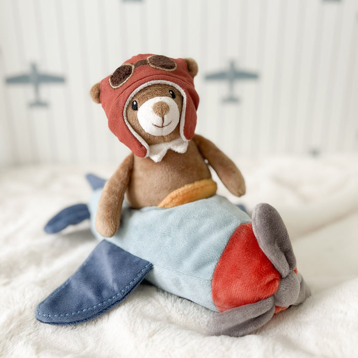 Stuffed teddy bear dressed as an aviator sitting on a toy airplane with a soft, blurred airplane pattern in the background.