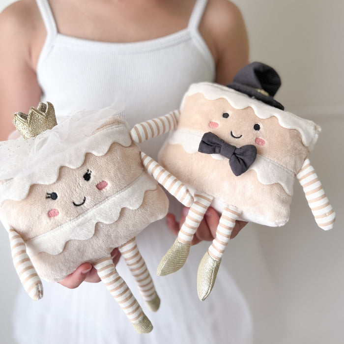 Two plush anthropomorphic cakes, one with a crown and veil, and the other with a top hat and bow tie, held by a person in a white tank top and dress.