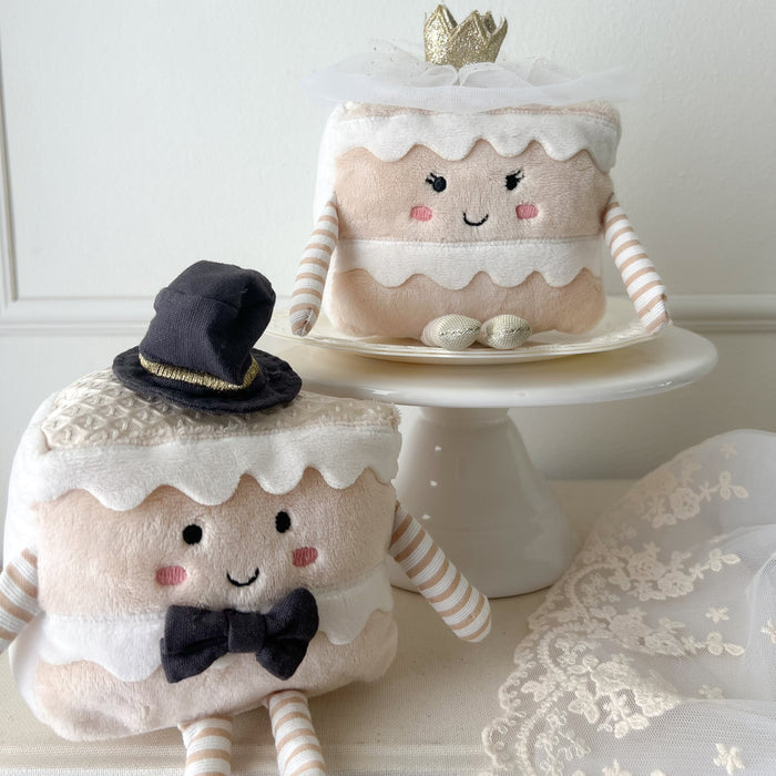 Two plush cake slice toys: one with a black top hat and bowtie, the other with a gold crown and tulle veil, displayed on a white cake stand with lace fabric nearby.