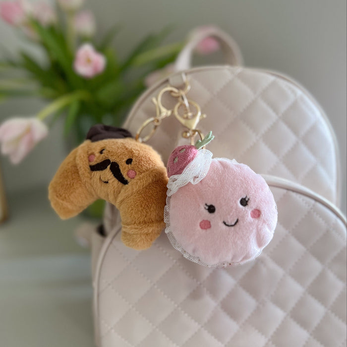 Two plush keychains, one dog-shaped and one macaron-shaped, attached to a white textured surface with flowers in the background.