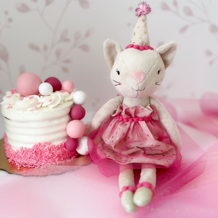 Stuffed cat wearing party hat and pink dress, next to frosted cake with pink sprinkles.