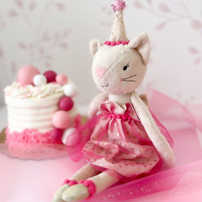 Stuffed cat toy wearing a party hat and pink dress next to a decorated cake.