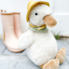Stuffed white duck wearing a yellow hat and checkered green scarf, sitting next to pink rain boots.
