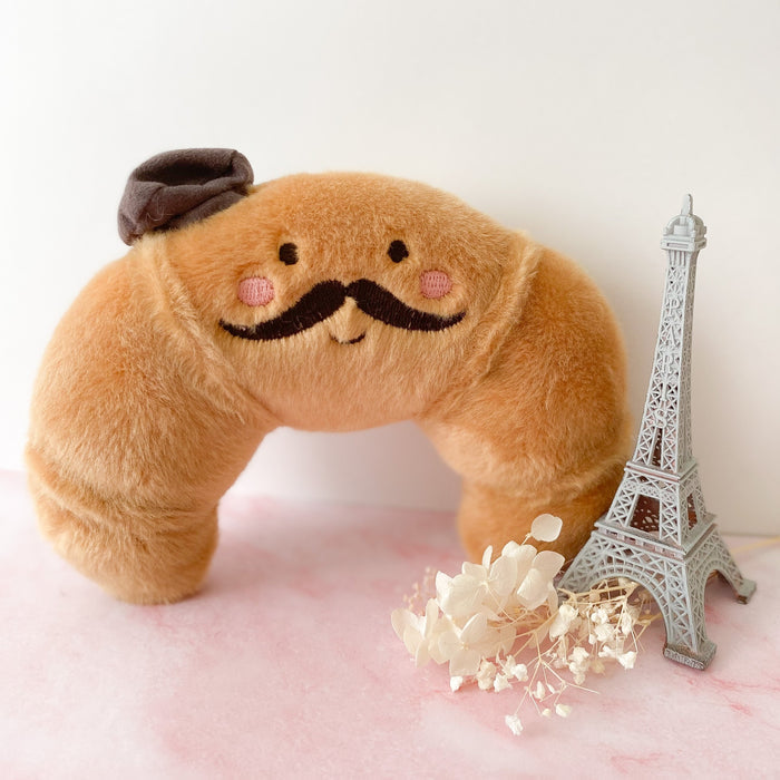 Plush croissant toy with a mustache, pink cheeks, and a brown hat, posed next to a miniature Eiffel Tower and dried white flowers on a pink marble surface.