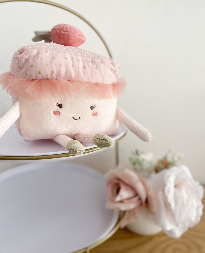 Plush pink cupcake toy with gold polka dots, tulle, and strawberry topping, displayed on a tiered tray with white plates and gold frame.