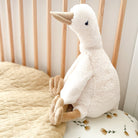 Stuffed goose sitting in a crib. The toy has a tan beak, legs, and feet, and a white plush body. A tan quilted blanket sits to the left, and a floral patterned pillow is underneath the toy.