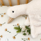 Close-up of a fluffy white goose plush toy on a floral print blanket with greenery.