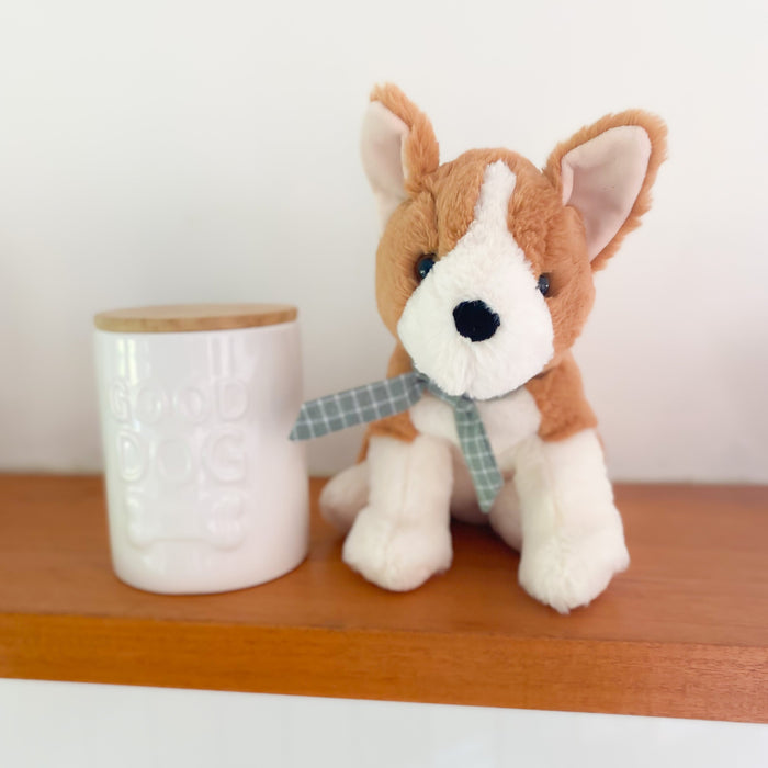 A stuffed dog with a plaid ribbon tie sitting next to a white ceramic jar with a wooden lid on a wooden shelf.