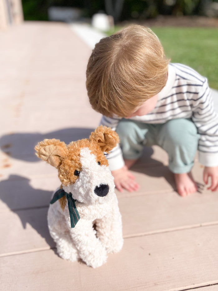 A toddler with blonde hair crouching next to a stuffed dog with a green ribbon on a wooden deck.