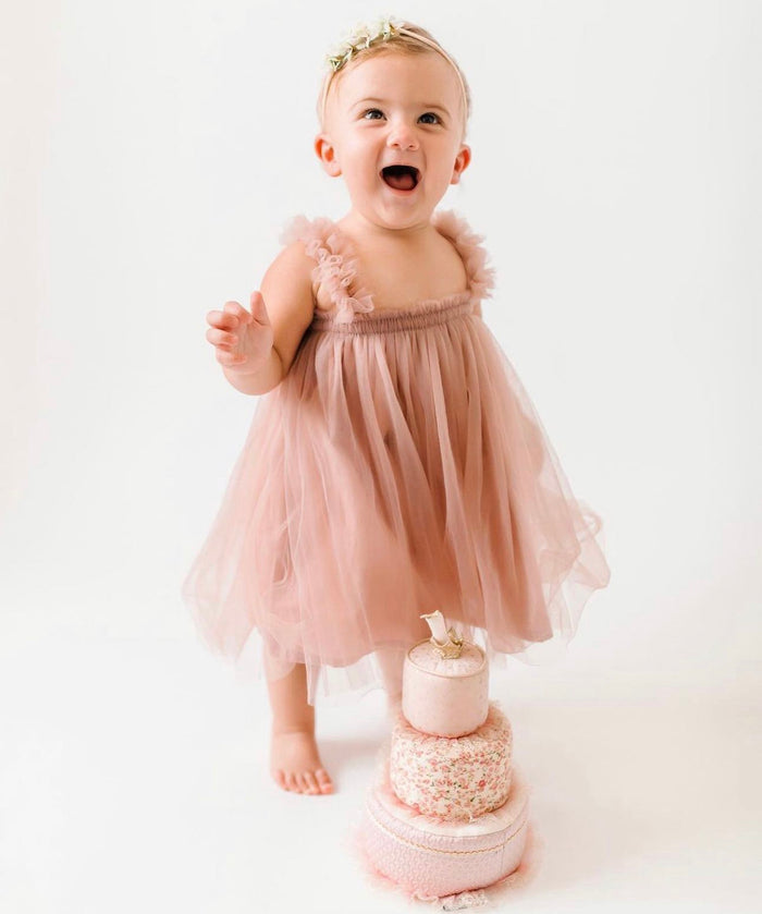 A young child in a pink tulle dress stands barefoot next to a three-tiered decorative cake.