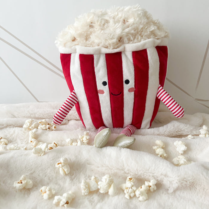 Plush popcorn container with red and white stripes, arms, smile, and popcorn surrounding it on a white blanket.