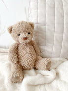 Close-up of a plush, light brown teddy bear sitting on a white blanket with a white, striped cushion in the background.