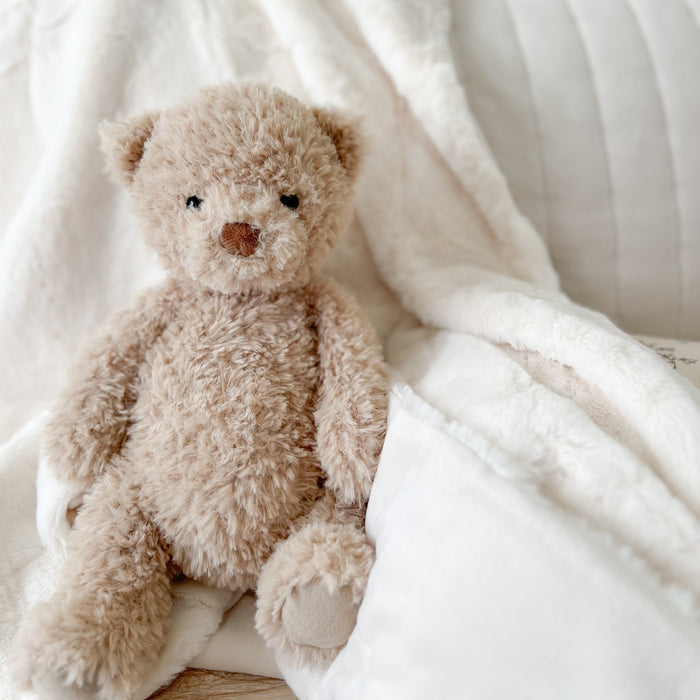 Tan teddy bear sitting on a soft white blanket.