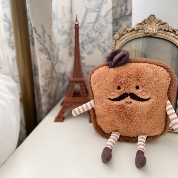 Plush toast character with a mustache, beret, striped limbs, and a miniature Eiffel Tower in the background.